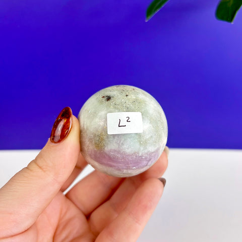 Polished pink tourmaline sphere with white and grey mineral inclusions held in a hand against a blue background.
