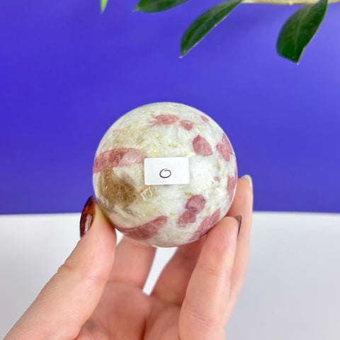 Polished pink tourmaline sphere with white and brown mineral inclusions held in a hand against a blue background.