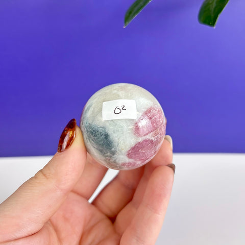 Polished pink and grey tourmaline crystal sphere held in hand against a purple and white background.