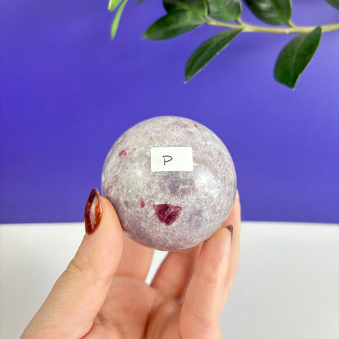 Polished pink tourmaline sphere with natural inclusions held in a hand against a purple and white background.