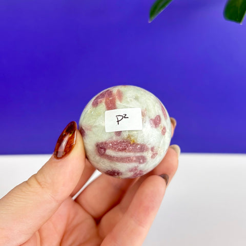 Polished pink tourmaline sphere with white quartz inclusions held in a hand against a blue background.
