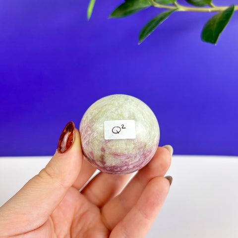 Hand holding a polished pink and pale green tourmaline sphere with a Q2 label against a blue background.