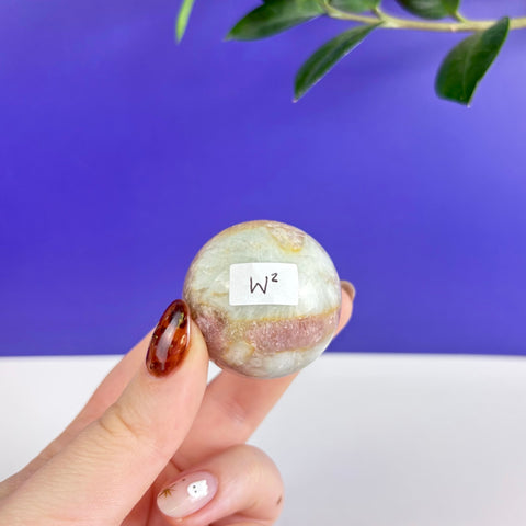 Polished pink tourmaline sphere with white and tan mineral inclusions held between fingers.