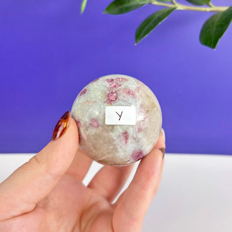 Polished pink tourmaline sphere with white quartz inclusions held in a hand against a purple background.