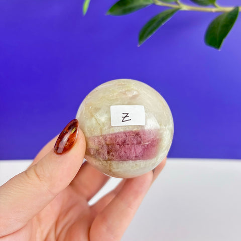 Polished petrifying pink tourmaline sphere with a vibrant magenta inclusion held in a hand against a blue background.