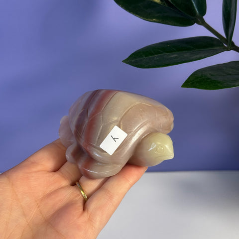 Pink banded onyx turtle carving, held in hand, with a light purple background.