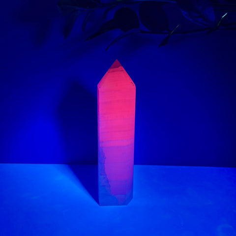 Polished pink calcite crystal tower glowing with a vibrant neon orange hue under UV light against a dark blue background.