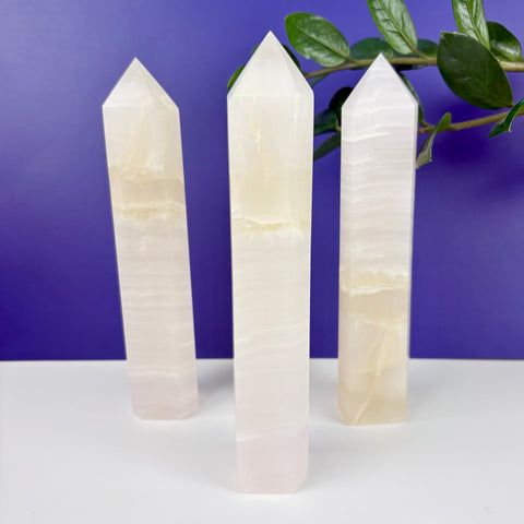 Three polished pink calcite crystal towers with banded textures standing on a white surface against a purple background.