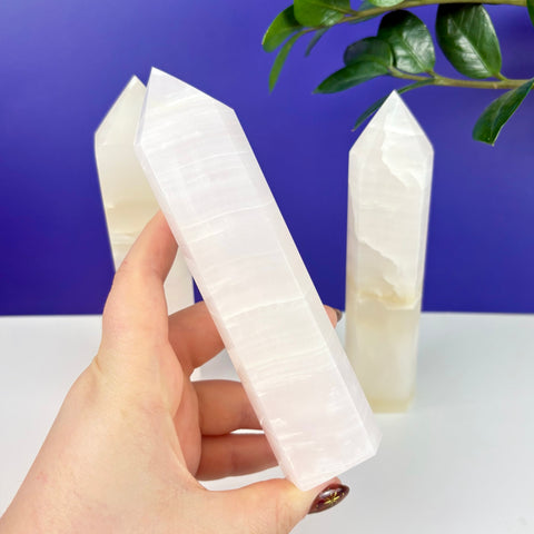 Hand holding a translucent, pale pink calcite crystal tower with smooth, polished facets against a blue background.