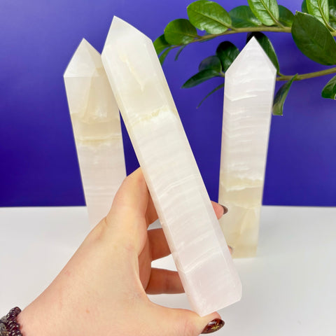 Hand holding a smooth, translucent pink calcite crystal tower with two other towers standing on a white surface.