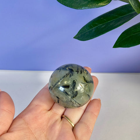 Hand holding a polished green prehnite sphere with dark tourmaline inclusions.