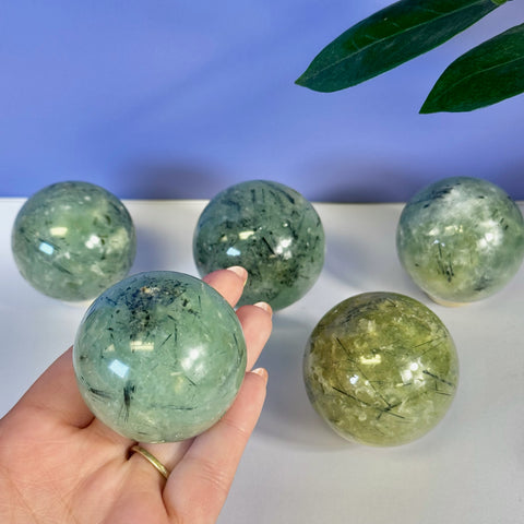 Hand holding a polished green Prehnite sphere with black tourmaline inclusions. Four more spheres are visible in the background.