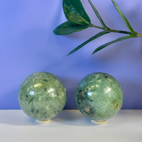 Two polished green Prehnite spheres with black tourmaline inclusions on white stands, against a purple wall.