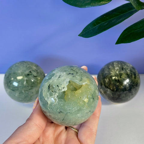 Hand holding a translucent green Prehnite sphere with a raw yellow crystal geode opening.
