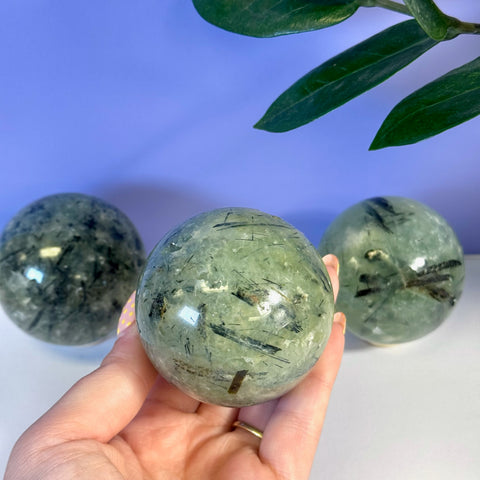 Hand holding a polished green Prehnite sphere with dark tourmaline inclusions. Two more spheres in background.
