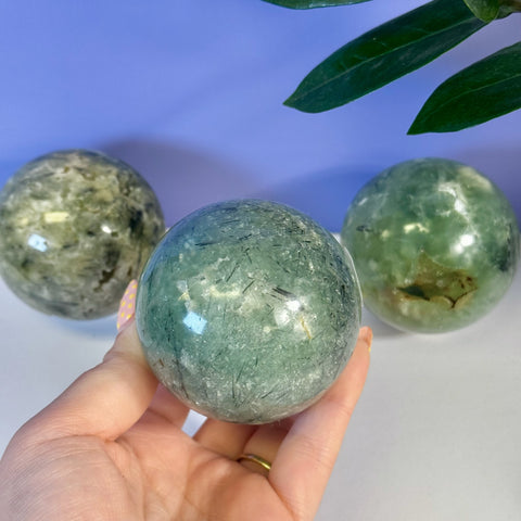 Hand holding a polished, translucent green Prehnite sphere with black tourmaline inclusions. Two other spheres in background.