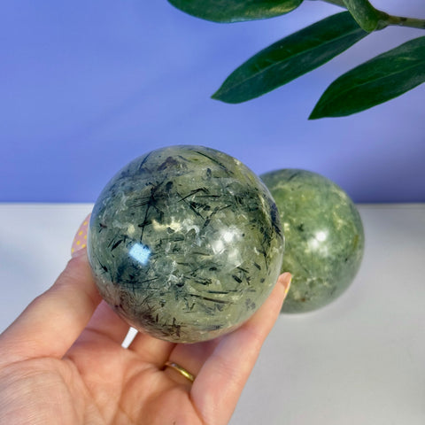 Hand holding a polished green Prehnite sphere with black tourmaline inclusions.