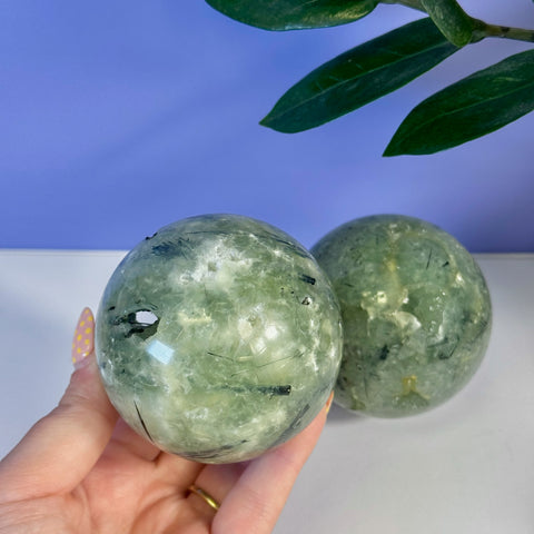 Smooth, polished green prehnite sphere with black tourmaline inclusions, held in hand.