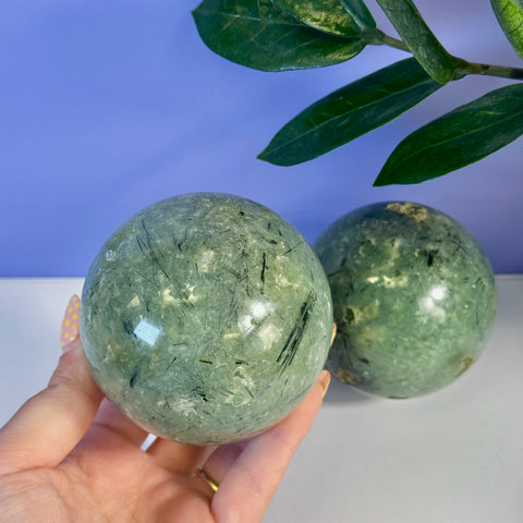 Smooth, polished green Prehnite sphere with black tourmaline inclusions held in hand, another sphere in background.