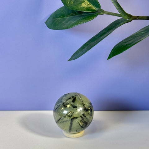 Smooth green Prehnite sphere with dark inclusions on a stand, against a purple wall with a plant.