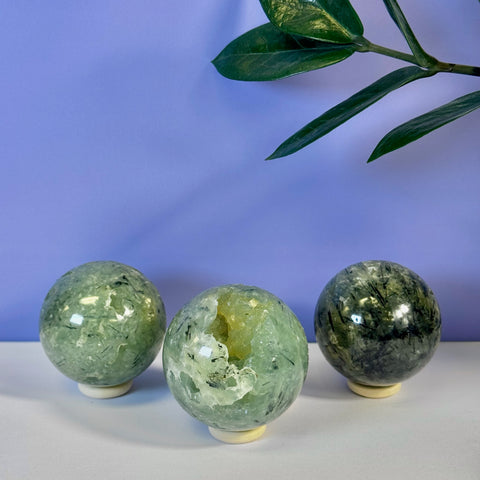 Three green Prehnite spheres, one with a yellow druzy geode, on a white surface with a purple background.