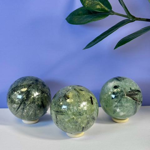 Three polished green Prehnite spheres with black tourmaline inclusions on white stands.