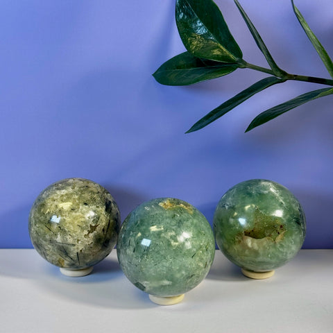 Three polished green Prehnite spheres with black inclusions, displayed on a white surface against a purple background.