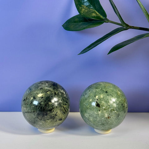 Two green Prehnite spheres with black tourmaline inclusions on white stands, against a purple wall.