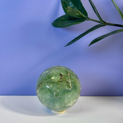 Smooth, polished green prehnite sphere with natural inclusions, on a white surface with a purple background.