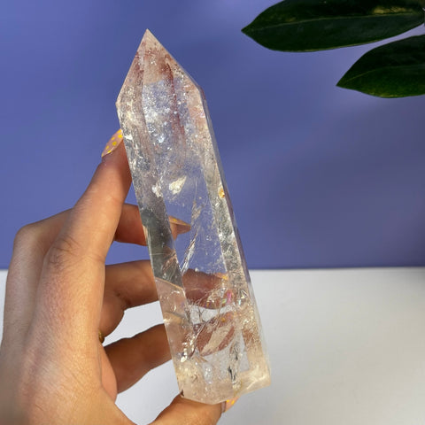 Clear Rainbow Pink Fire Azeztulite crystal tower with natural inclusions, held against a purple background.