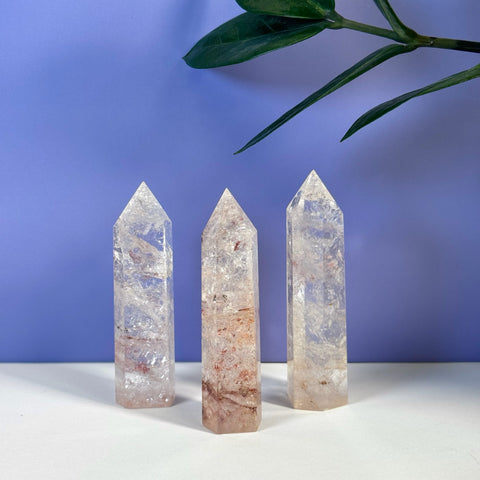 Three clear Rainbow Pink Fire Azeztulite crystal towers with reddish inclusions on a white surface.