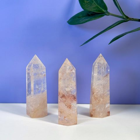 Three clear Rainbow Pink Fire Azeztulite crystal towers with reddish inclusions, against a purple background.