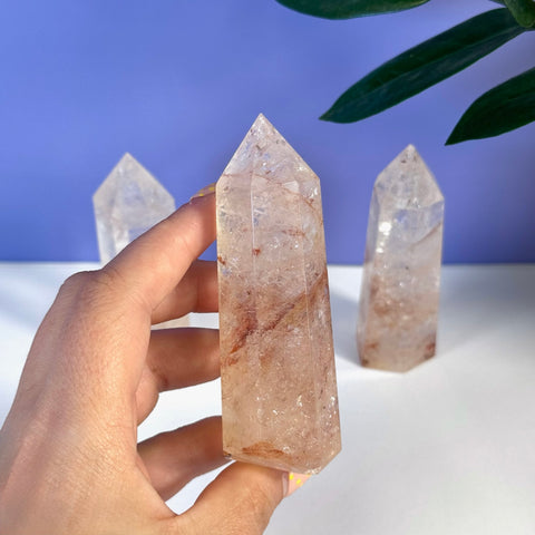 Hand holding a Rainbow Pink Fire Azeztulite crystal tower with red inclusions; two more towers in background.