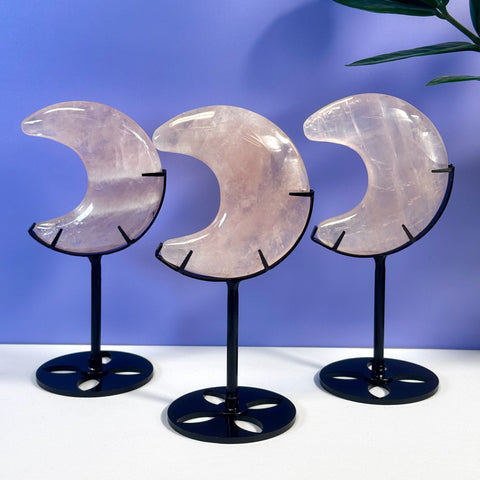 Three rose quartz crescent moons on black metal stands against a purple wall.