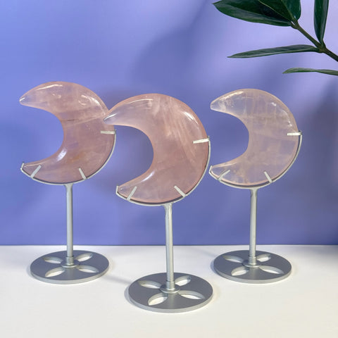 Three rose quartz crescent moons on silver stands against a purple background.