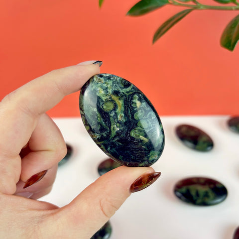 Polished oval Kambaba Jasper palm stone with dark green and black swirling patterns held between fingers.