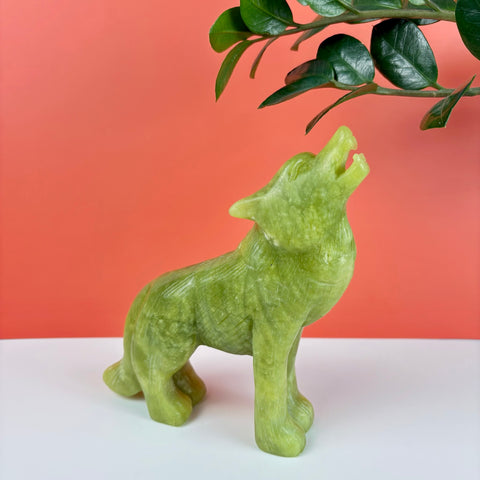 Hand-carved light green serpentine stone wolf figurine howling, set against a coral and white background.