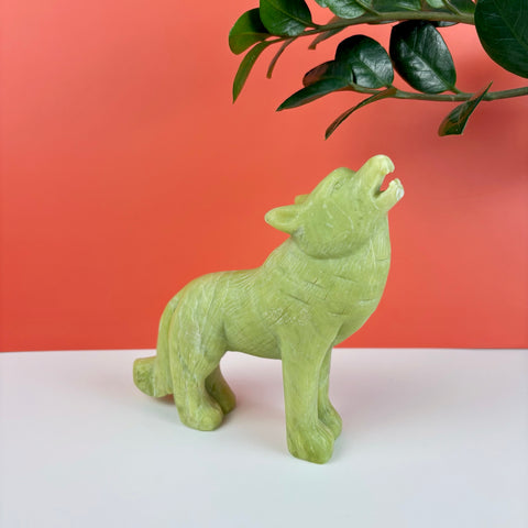 Hand-carved light green serpentine stone wolf figurine howling, set against a vibrant coral and white background.