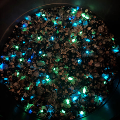 Assorted polished protection crystals and stones glowing with bright blue and green light in a dark bowl.