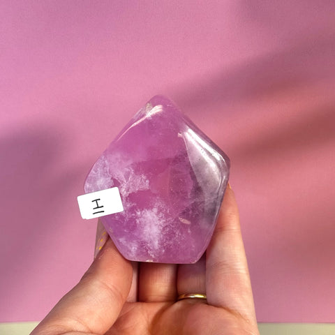 Polished pink aragonite free form crystal with white inclusions, held against a pink background.