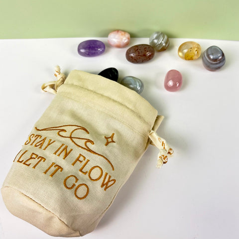 Cream canvas drawstring pouch with gold embroidery and ruffled top, surrounded by assorted polished crystals.