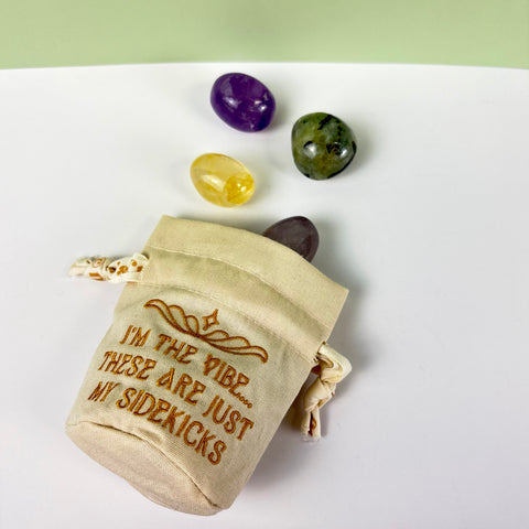 Small beige canvas drawstring pouch with gold embroidery and three polished crystals on a white surface.