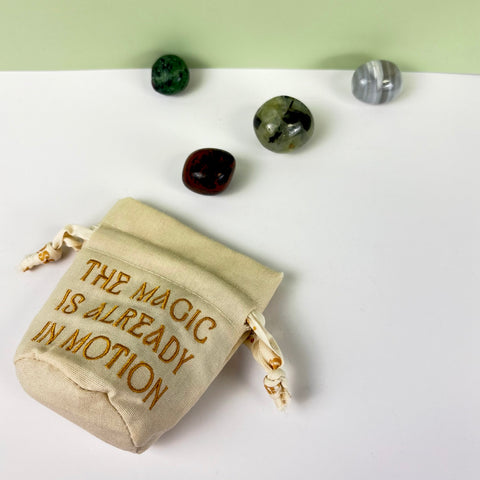 Small cream canvas drawstring pouch with gold embroidery and four polished crystals on a white surface.