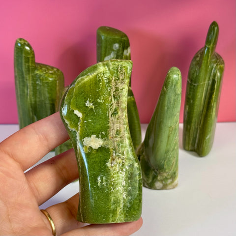 Hand holding a polished, vibrant green tremolite free form with white veins, against a pink background.