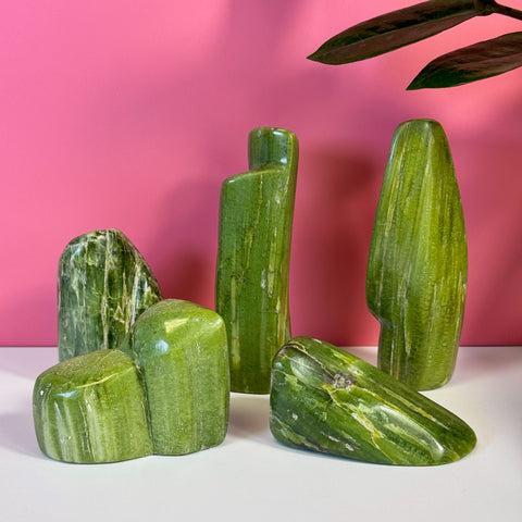 Five polished green Tremolite free forms with white striations on a white surface, pink background.
