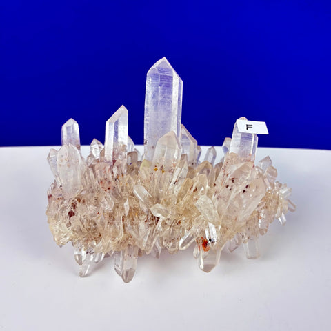 Cluster of clear Himalayan quartz crystals with reddish inclusions on a white surface against a blue background.