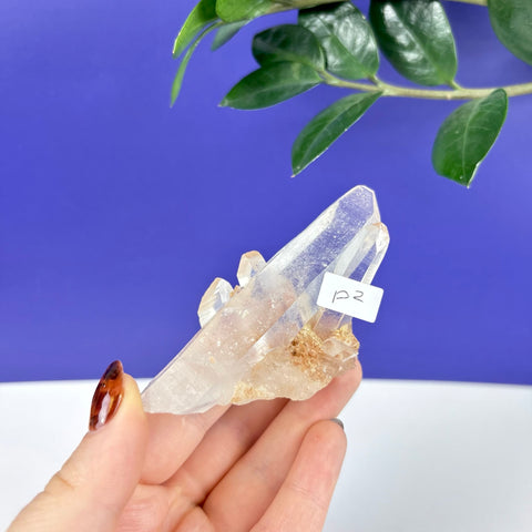 Hand holding a clear, pointed Himalayan quartz crystal cluster with earthy mineral inclusions against a blue background.