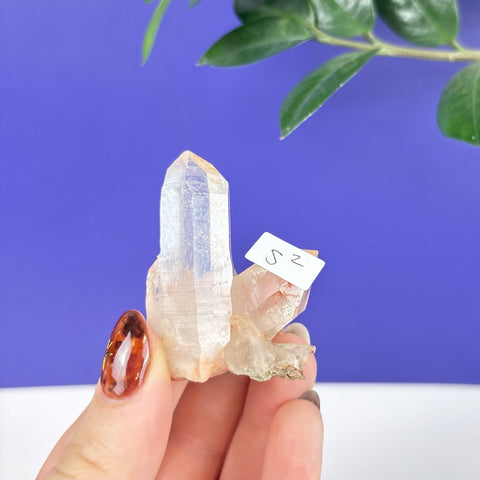 Clear Himalayan quartz crystal cluster with a small S2 label, held against a vibrant purple background.